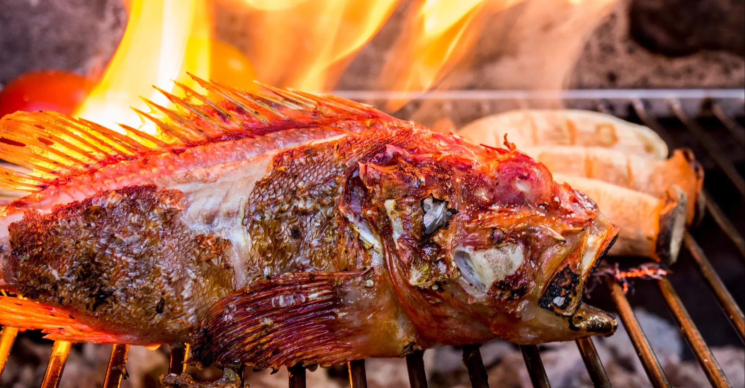 Ganzer Fisch auf dem Holzkohlegrill, dahinter die Flammen